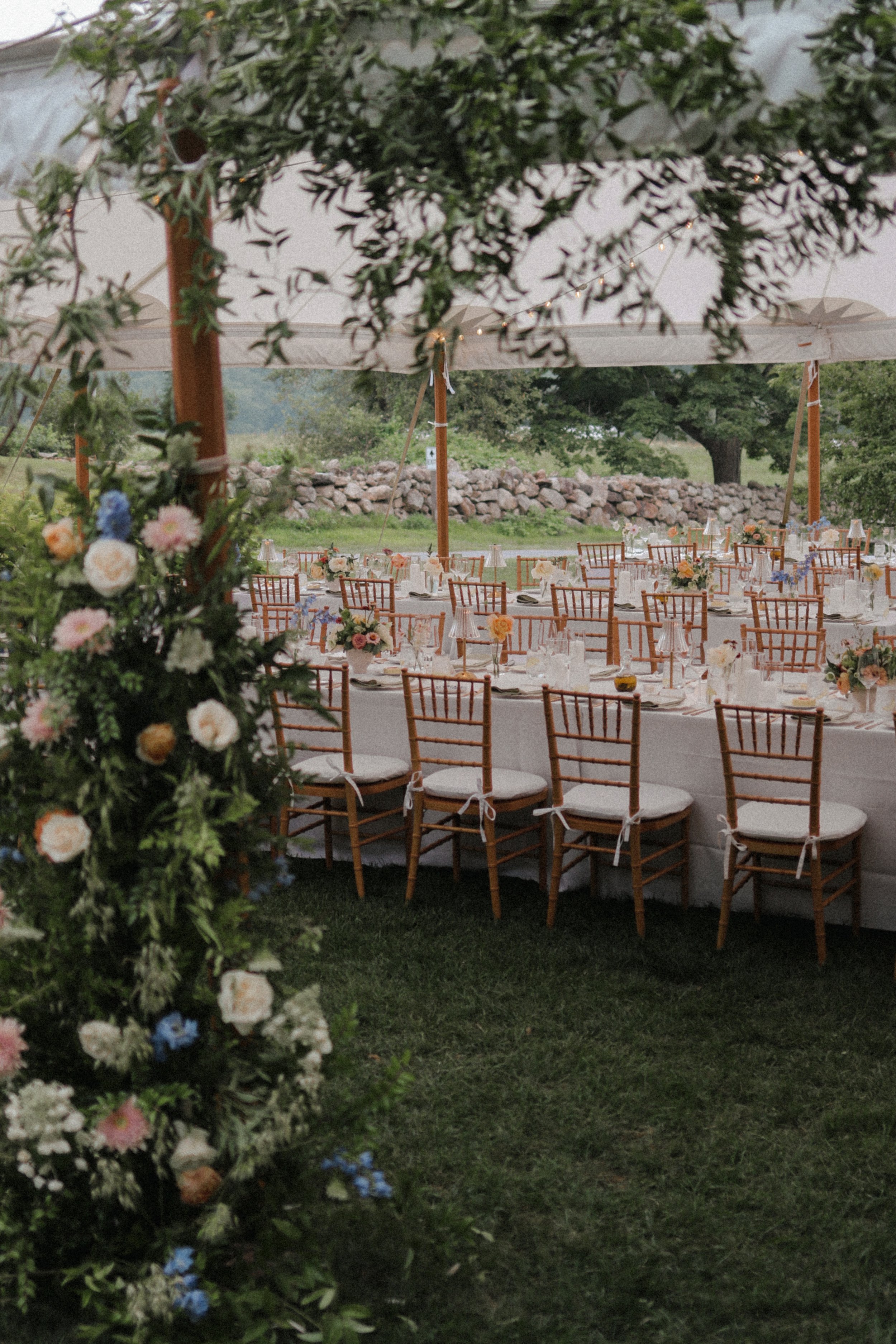 Decorated outdoor dining area under a large white canopy with a long table, floral centerpieces, and wooden chairs, set in a grassy area with trees and rocks in the background.
